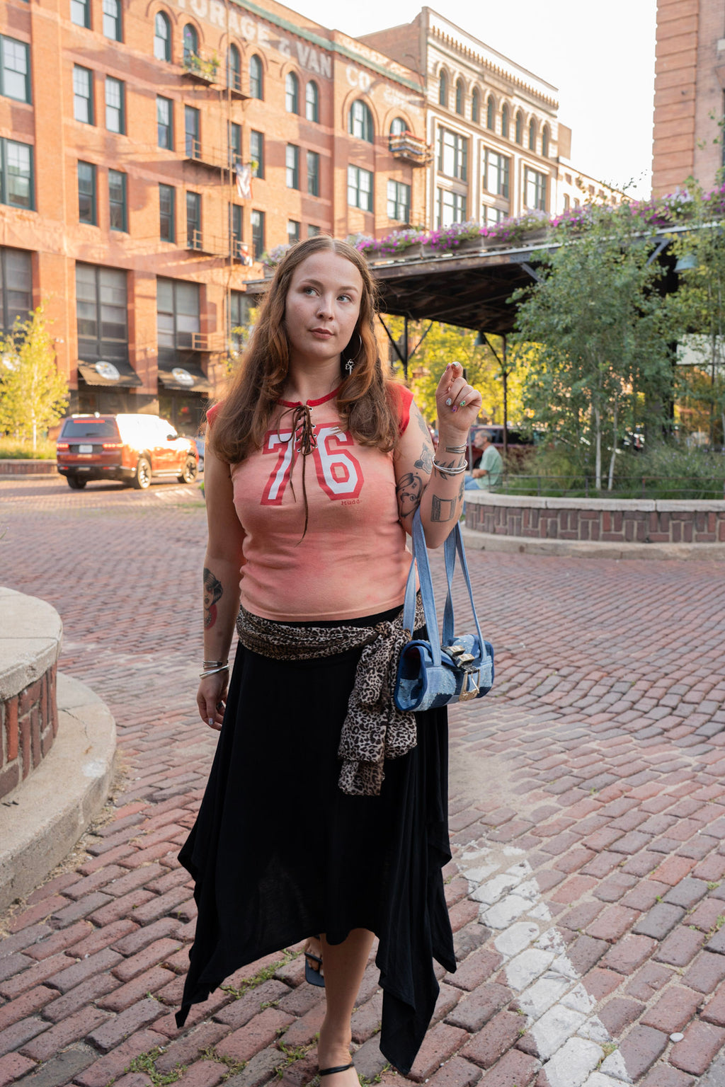 A woman stands on a brick street in front of brick buildings, wearing a pink cropped tank top with '76' printed on it, a long black skirt, and carrying a blue shoulder bag.