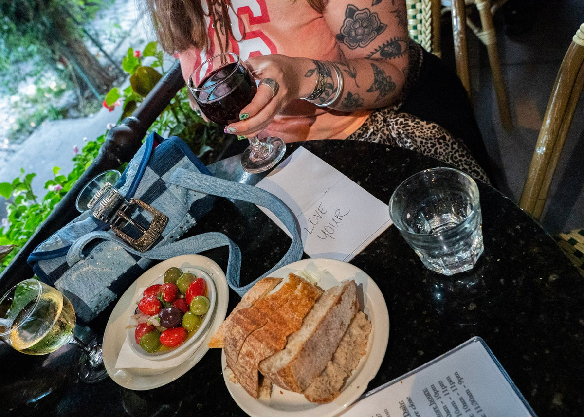 A person seated at a restaurant table holding a dark wine glass, with plates of bread, a small plate of fruit, a water glass, and a blue denim bag on a dark tabletop.