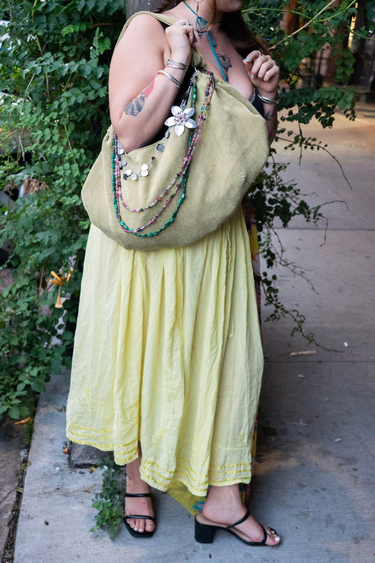A woman wearing a flowing pale yellow maxi dress and black sandals, carrying a large olive green shoulder bag decorated with white flowers and layered beaded necklaces, standing outdoors on a concrete path with green foliage on the left.