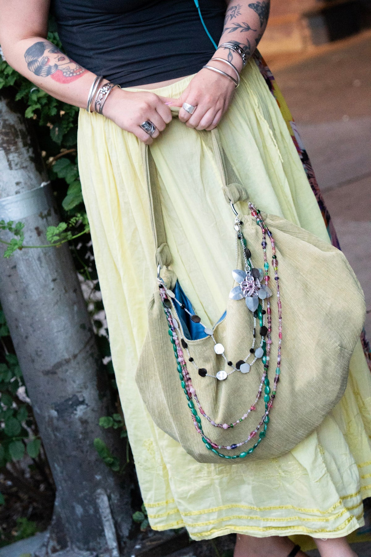A person outdoors wearing a pale yellow skirt and holding a large, draped sage green fabric tote bag adorned with colorful beaded necklaces and charms.