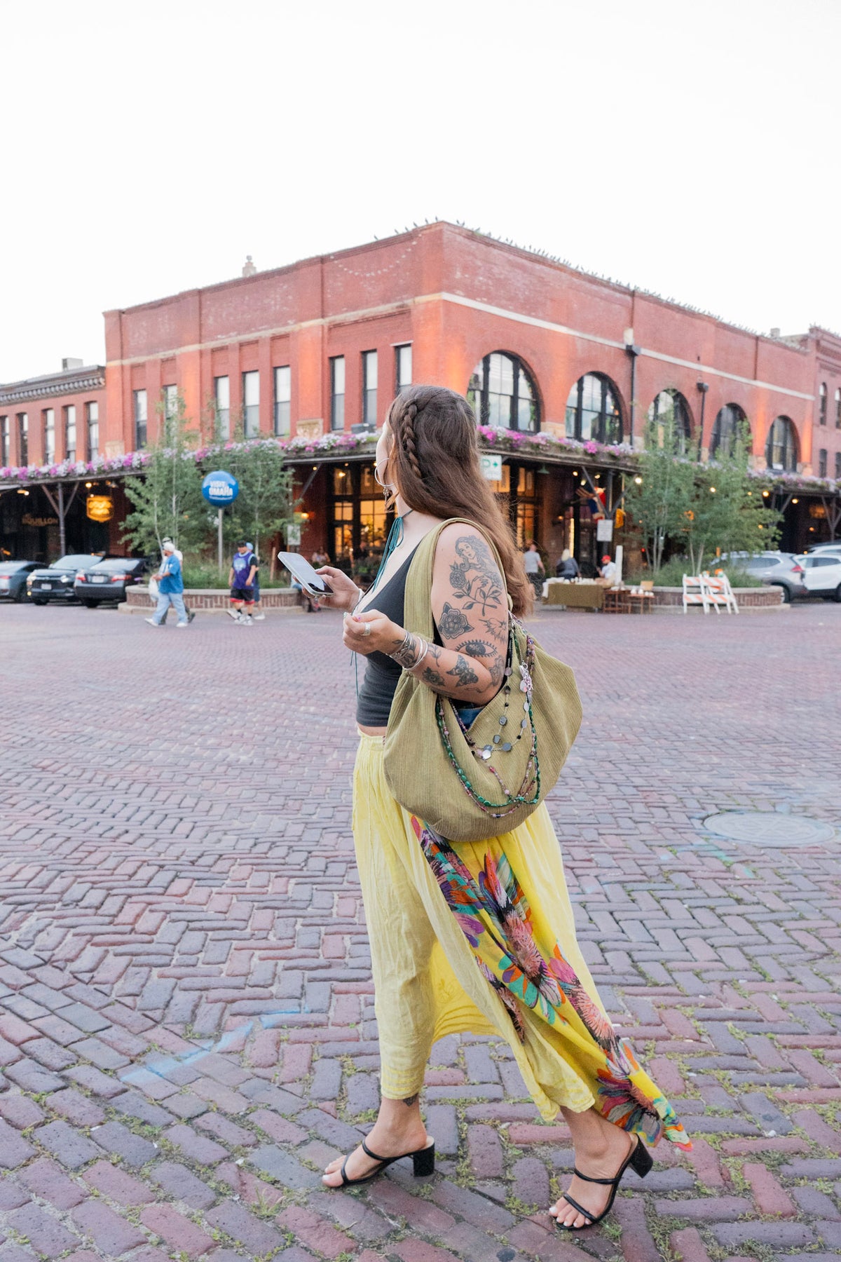 A woman with tattoos walks on a brick plaza wearing a black top, a flowing yellow skirt with a multicolor floral print, and carrying a large olive green shoulder bag.