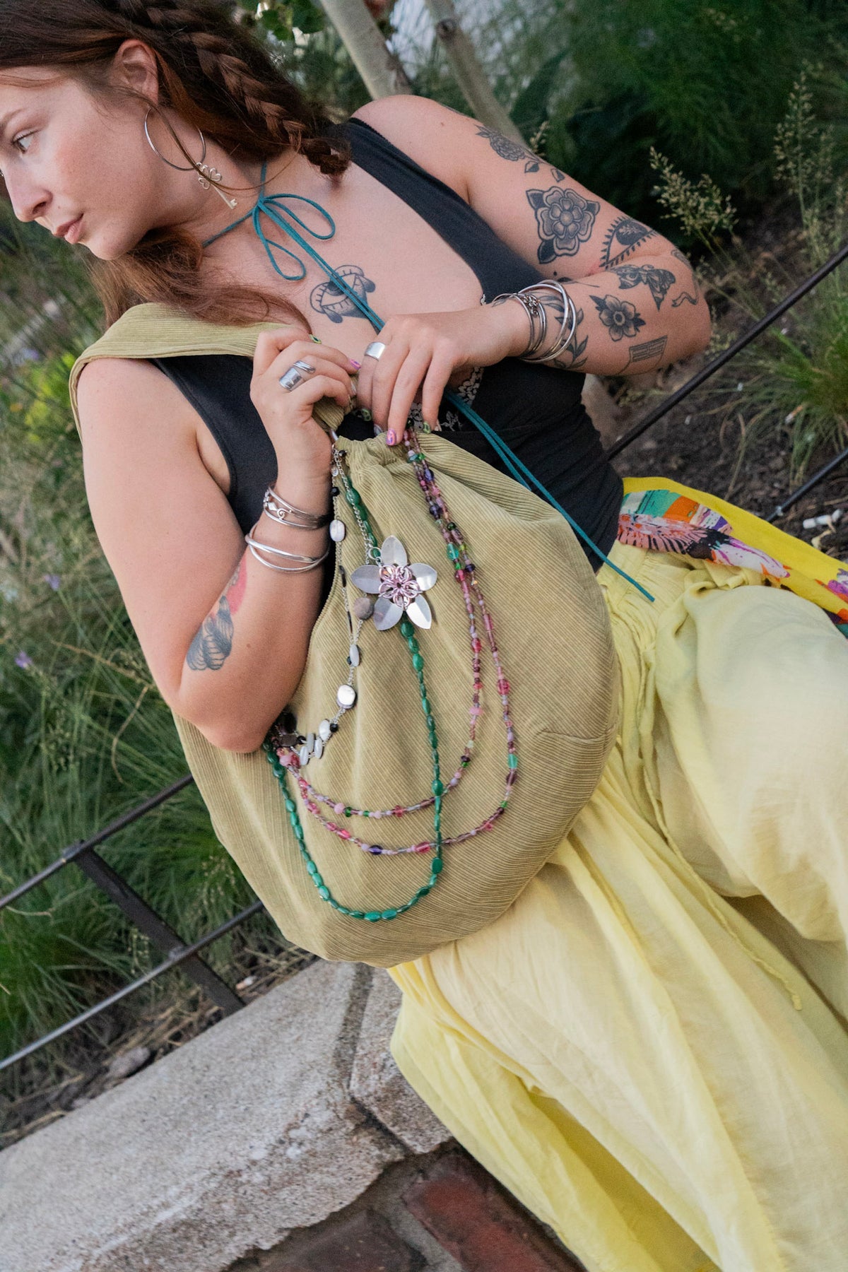 A woman outdoors wearing a long yellow skirt and black top, holding a large olive-brown fabric drawstring bag decorated with colorful beaded necklaces and a silver flower charm.