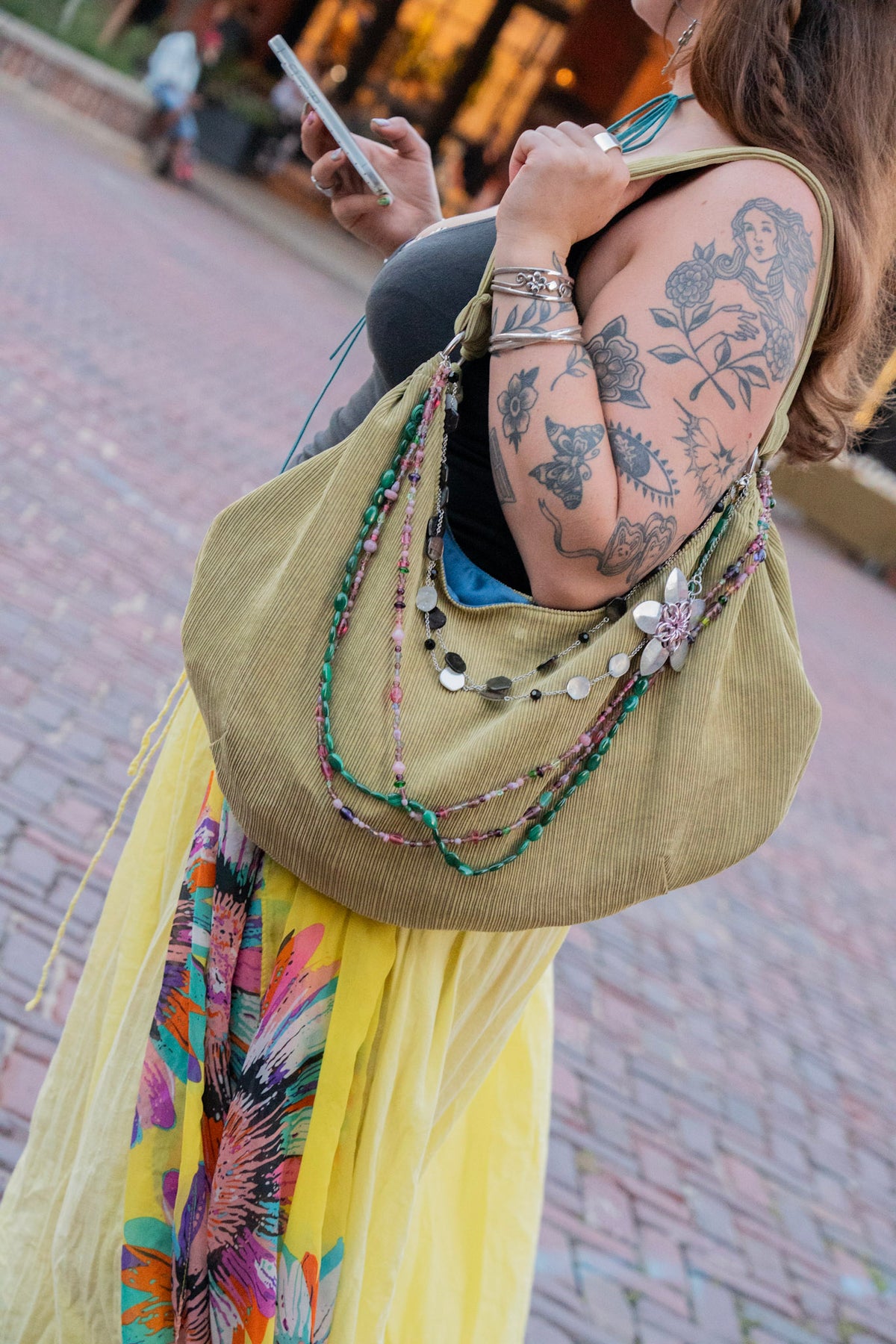 A woman with arm tattoos carries a large olive green corduroy hobo shoulder bag decorated with colorful beaded strands and silver coin accents. She is also wearing a bright yellow skirt with a colorful floral print.