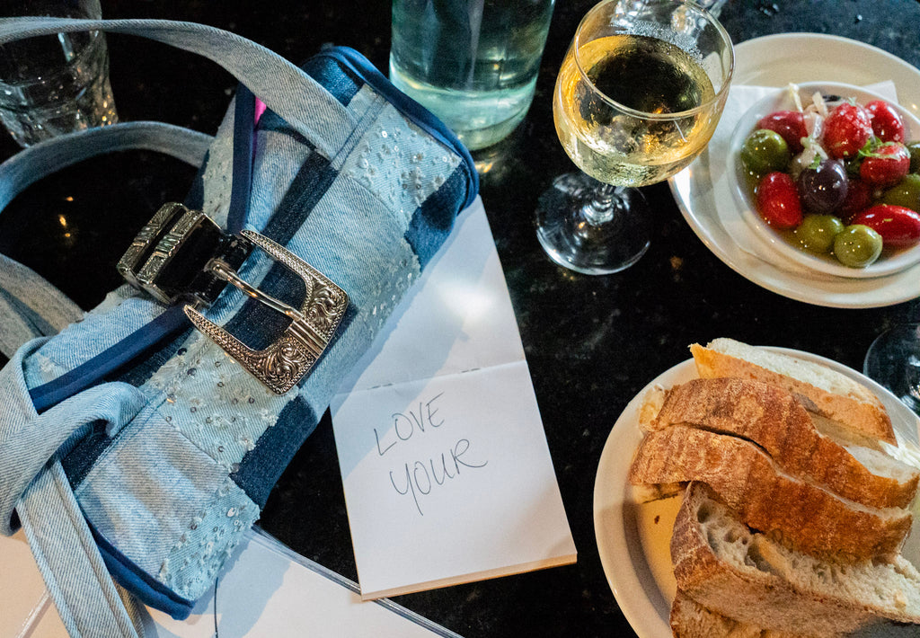 A close-up lifestyle shot of a light blue denim handbag with a silver buckle, placed on a dark countertop next to a handwritten note that says 'LOVE YOUR', a glass of white wine, a plate of sliced bread, and a plate of olives.
